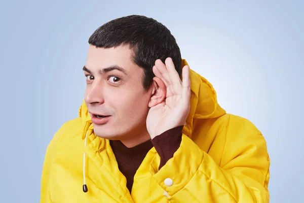 Curious brunet male model tries to overhear something, keeps hand on ear, listens gossips in next room, wears yellow anorak, isolated over blue background. Young man listens something attentively — Stock Photo, Image