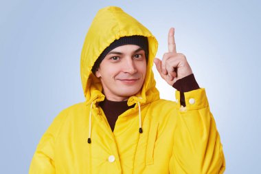Akıllı yakışıklı adam yükseltir ön plana parlak bir fikir, giyer sarı yağmurluk, alır gibi parmak üzerinde mavi arka plan izole mutlu neşeli ifade vardır. Şık giysiler jestleri Studio içinde genç erkek