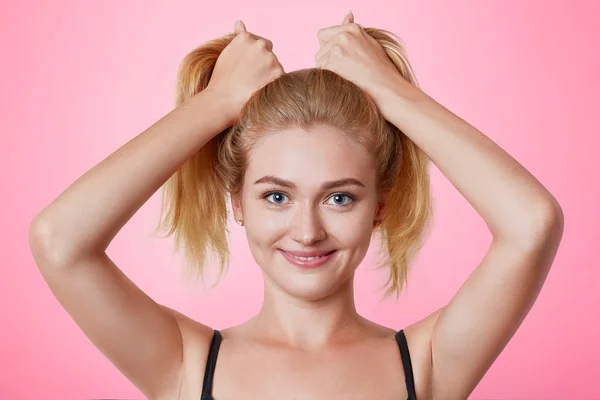 Foto de divertida modelo femenina alegre hace dos colas de caballo de pelo, se para frente al espejo, piensa qué peinado hacer como ir de fiesta con amigos. Preparación para un evento especial. Divertirse — Foto de Stock