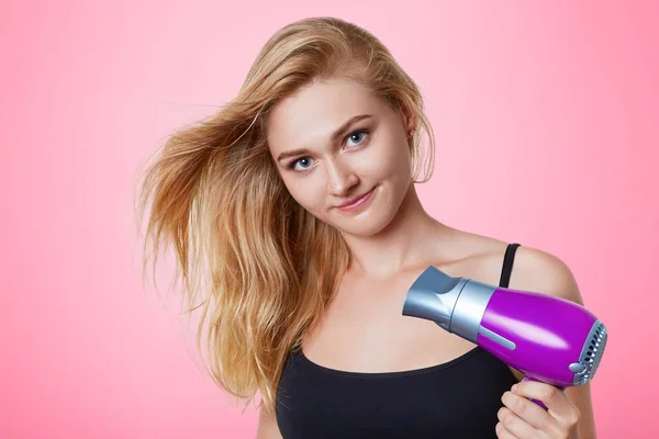 Concepto de cuidado del cabello. Hermosa mujer utiliza secador de pelo, golpes secan su cabello, se prepara para la fecha con novio guapo. Bastante joven rubia hembra se preocupa por la apariencia, aislado sobre fondo rosa — Foto de Stock