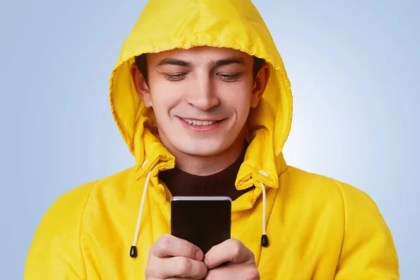 Ευτυχής θετική αρσενικό φοράει κίτρινο μπουφάν και κουκούλα, τύποι μηνυμάτων για το έξυπνο τηλέφωνο, χρησιμοποιεί δωρεάν σύνδεση στο internet. Όμορφος άνδρας χρησιμοποιεί σύγχρονες elecrtonic συσκευή για την online επικοινωνία. Τεχνολογία έννοια — Φωτογραφία Αρχείου