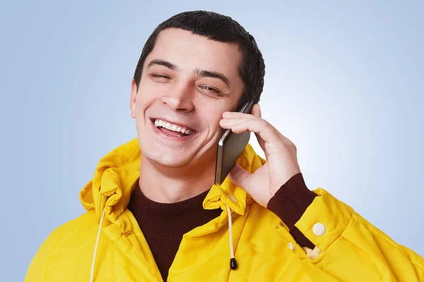 年轻快乐帅气的男人有电话交谈, 和最好的朋友谈话, 用高兴的表情讨论一些事情, 高兴地听他说, 穿黄色的皮猴孤立在蓝色背景上。技术理念 — 图库照片