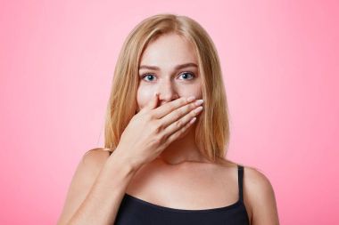 Hoş görünümlü hafif saçlı erkek kayıtsızca giyinmiş, pembe studio arka plan üzerinde sessiz ve değil sır için çalışır bir izole kapakları el ile mouth. İnsanlar, güzellik, vücut dili kavramı