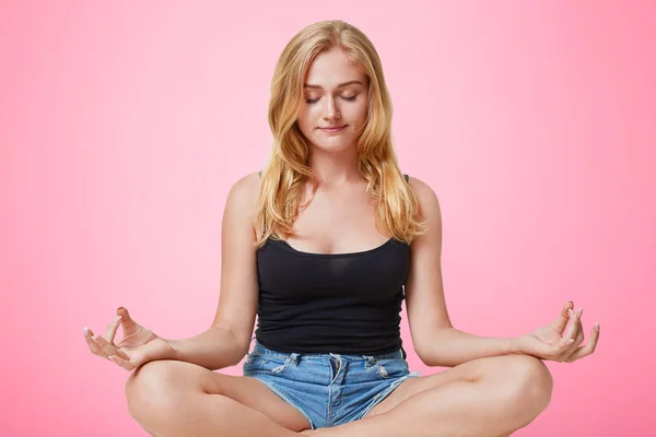 Портрет красивої жіночої моделі сидить в позі йоги, медитує на підлозі, закриває очі, тримає ноги схрещеними, ізольованими на рожевому тлі. Красива жінка практикує йогу в приміщенні. Концепція фітнесу та спокою — стокове фото