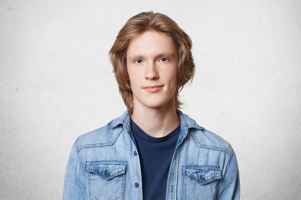 Agradável olhar elegante macho com penteado na moda, veste camisa jeans, olha a sério diretamente na câmera, isolado sobre fundo de concreto branco. Estudante masculino confiante em seu conhecimento — Fotografia de Stock