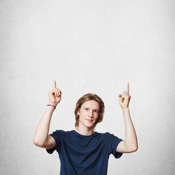 Attractive young European male with trendy hairdo, wears casual t shirt, indicates upwards as advertizes something, isolated over white concrete wall. Vertical portrait. Advertising concept. Stock Picture
