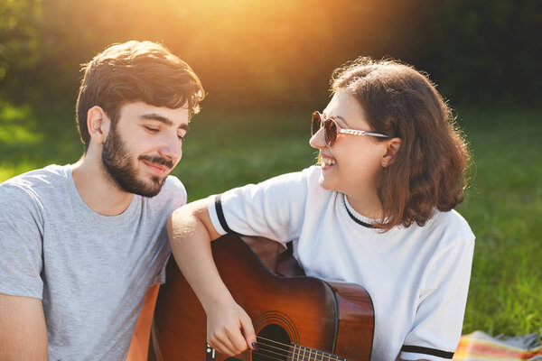 Outdoor shot of cheerful lovely couple spend free time on nature, play guitar and sign songs, look with love and cheerful expression at each other, enjoy beautiful nature and togetherness