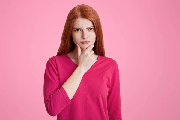 Isolerade skott av attraktiva röda haired kvinna håller hakan med hand, tycker om hennes problem, har trumpen uttryck, isolerade över rosa bakgrund. Tankeväckande vacker ingefära kvinna inomhus. — Stockfoto