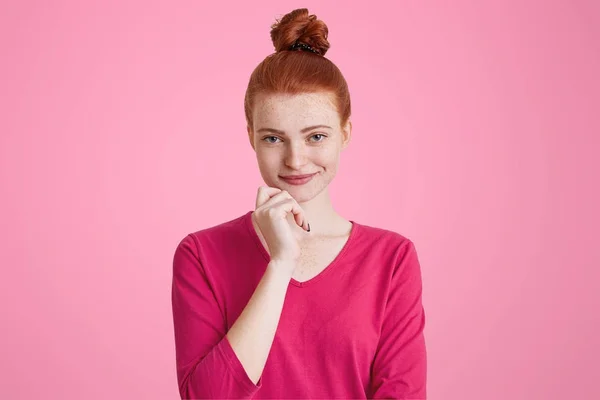 Hermosa modelo femenina con la piel pecosa tiene una sonrisa encantadora, el pelo rojo atado en el moño, mantiene la mano en la barbilla, aislado sobre el fondo rosa. Mujer europea está en el estudio. Concepto de emociones agradables —  Fotos de Stock