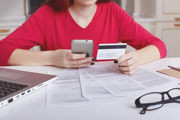 Cropped shot of unrecognizable female model in red sweater sits at working table surrounded with papers and laptop computer, uses cell phone for paying bill online with credit card. Ecommerce concept — Stock Photo, Image