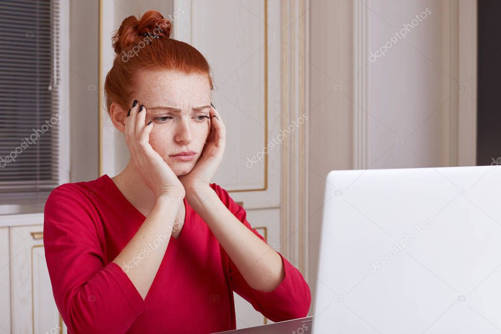 Puzzled young female student prepares for exams during last day, reads material from laptop computer, tries to remember something, sits over home kitchen interior. Education and technology concept