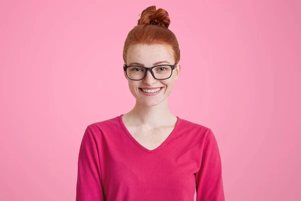 Foto aislada de mujer jengibre feliz en suéter rosa, lleva gafas cuadradas, tiene el pelo rojo atado en nudo, piel pecosa, sonrisa agradable, aislado sobre fondo rosa. Alegre estudiante mujer interior — Foto de Stock