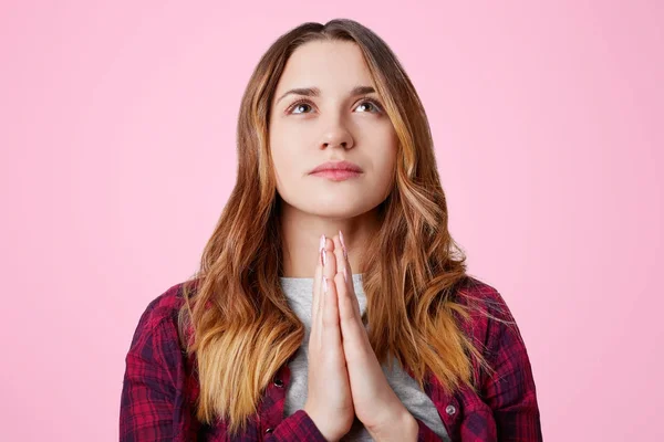 Retrato de fiel hembra adorable mantiene las manos juntas, pide a Dios bienestar y salud, se ve con suerte hacia arriba, aislado sobre fondo de estudio rosa. Mujer atractiva disfruta de un ambiente tranquilo — Foto de Stock