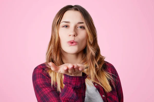 Bonitos golpes femeninos jóvenes besan a cámara, vestida con camisa casual a cuadros, expresa su amor y simpatía, aislada sobre fondo rosa. Linda hermosa joven mujer hace aire beso interior . — Foto de Stock