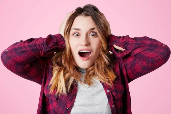 Photo of amazed excited female opens mouth and keeps hands on head, screams in excitement, doesn`t expect to hear some news, poses against pink studio background. Excitement and amazement concept