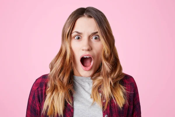 Sorprendido estudiante mujer estupefacta, abre la boca ampliamente, sorprendido de saber que mañana es el examen final, lleva camisa a cuadros, aislado sobre fondo rosa. Concepto de emociones humanas negativas — Foto de Stock