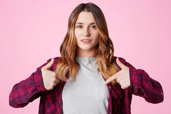 Mujer joven hermosa positiva con sonrisa agradable, indica en espacio de copia en blanco de la ropa para su diseño o contenido publicitario, aislado sobre fondo rosa. Concepto de ropa. Disparo horizontal — Foto de Stock