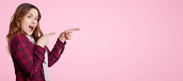 Emocionada hermosa mujer se para de lado, lleva chemise, puntos con los dedos delanteros a un lado en el espacio de copia en blanco para su anuncio o texto promocional. Alegre modelo femenino posa en estudio rosa . — Foto de Stock