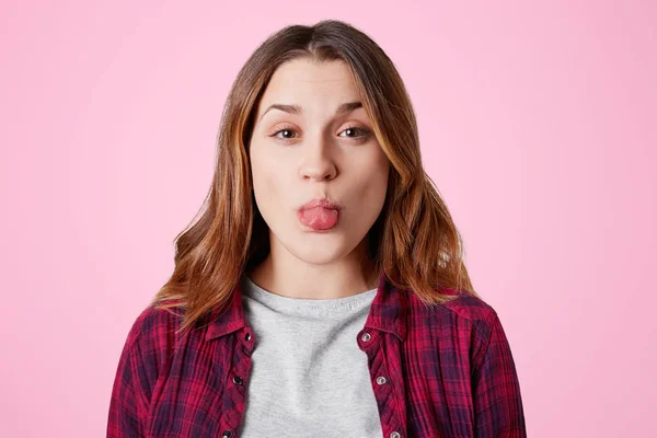 Divertida mujer bonita tontos en el interior, muestra la lengua, hace mueca, lleva chemise a cuadros, aislado sobre fondo de estudio rosa. Linda hembra joven se divierte, demuestra la lengua en el interior de la cámara — Foto de Stock