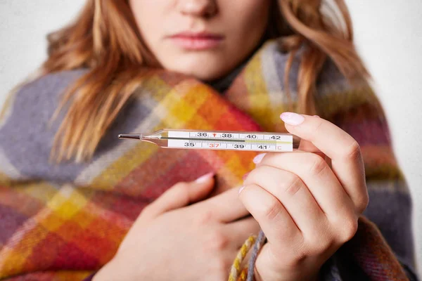 Περικοπεί shot από νεαρές τυλιγμένο σε ζεστό coverlet, κατέχει termometer που δείχνει υψηλή θερμοκρασία, έχει γρίπη, απομονώνονται σε φόντο λευκό στούντιο. Ανθυγιεινή η γυναίκα έχει κακό κρύο χειμώνα — Φωτογραφία Αρχείου