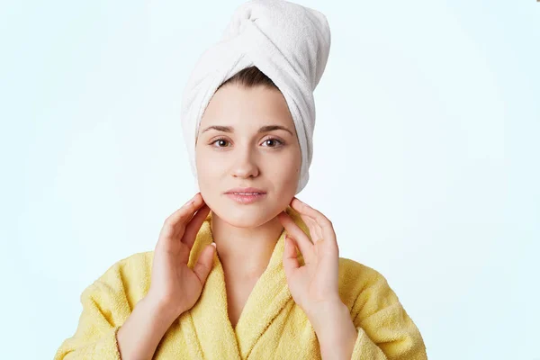 Adorable joven encantadora mujer en albornoz y toalla, tiene ducha, posa sobre fondo blanco, contenta de pasar tiempo en casa, tiene procedimiento de spa. Gente, higiene, belleza y concepto de salud —  Fotos de Stock