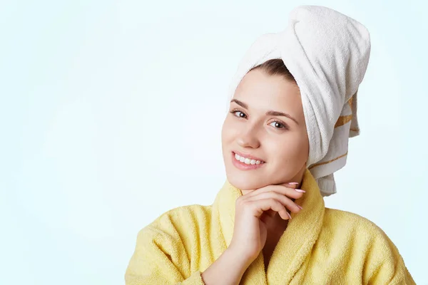 Horizontal shot of cheerful beautiful woman with bath towel on head dressed in yellow bathrobe, has just taken shower. Young attractive female being at home. Spa, bodycare and bathing concept — Stock Photo, Image