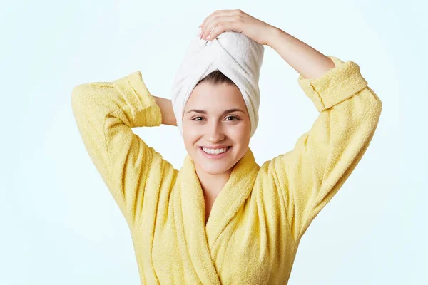 Glad beautiful female wears bathrobe and towel on head, glad to take shower, has appealing appearance, stands against white background. Relaxed cheerful woman after bathroom. Bathing concept — Stock Photo, Image
