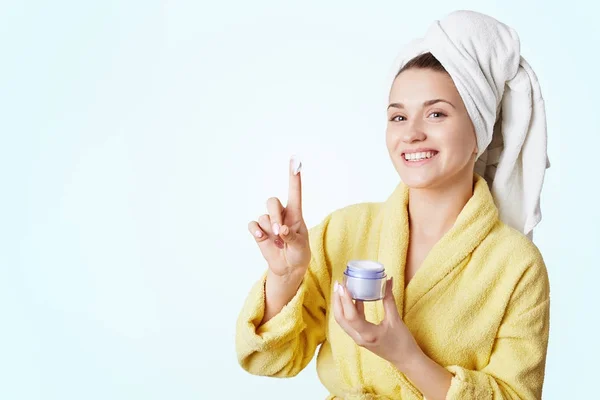 Happy smiling young woman wears bathrobe and towel, holds cream, advertizes its high quality and effect, guarantees soft skin after using it. Women, cosmetic, spa, beauty and skincare concept — Stock Photo, Image