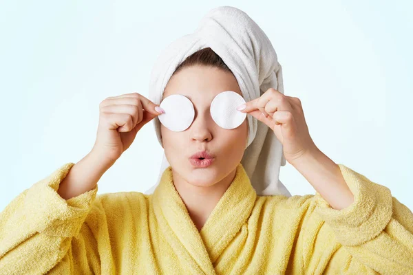 Funny woman foolishes after taking shower, wears towel and bathrobe, covers eyes with cotton wheels, going to apply cosmtics and do make up, isolated on white background. Beauty and hygiene concept — Stock Photo, Image