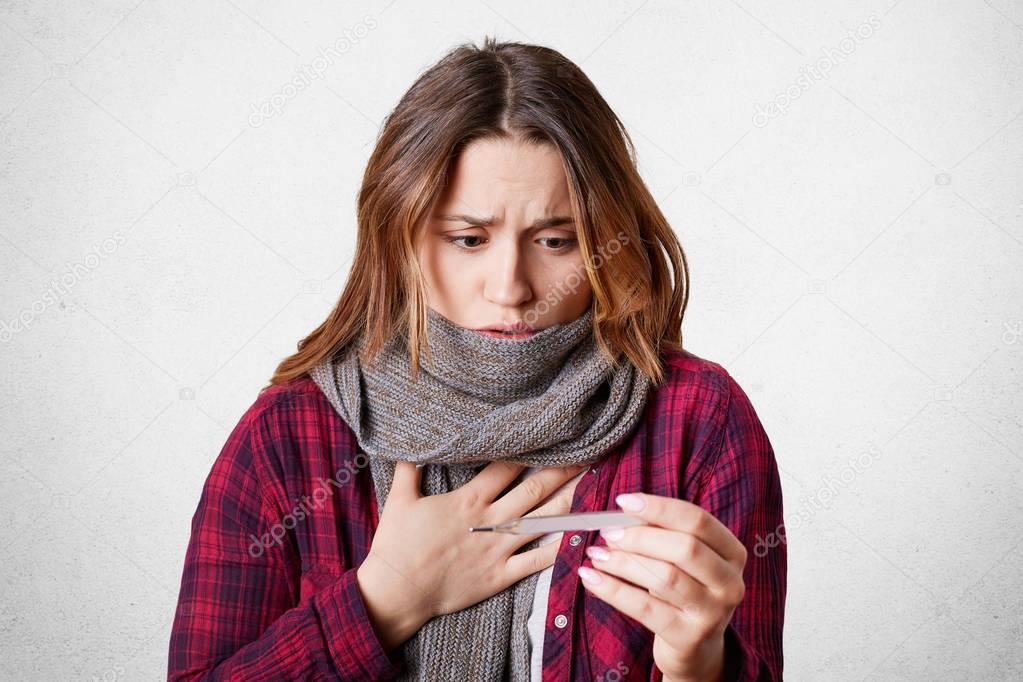 Sick beautiful woman looks at termometer, has high temperature, flu, wears warm scarf on neck as has sore throat, shocked, isolated over white background. People and seasonal disease concept