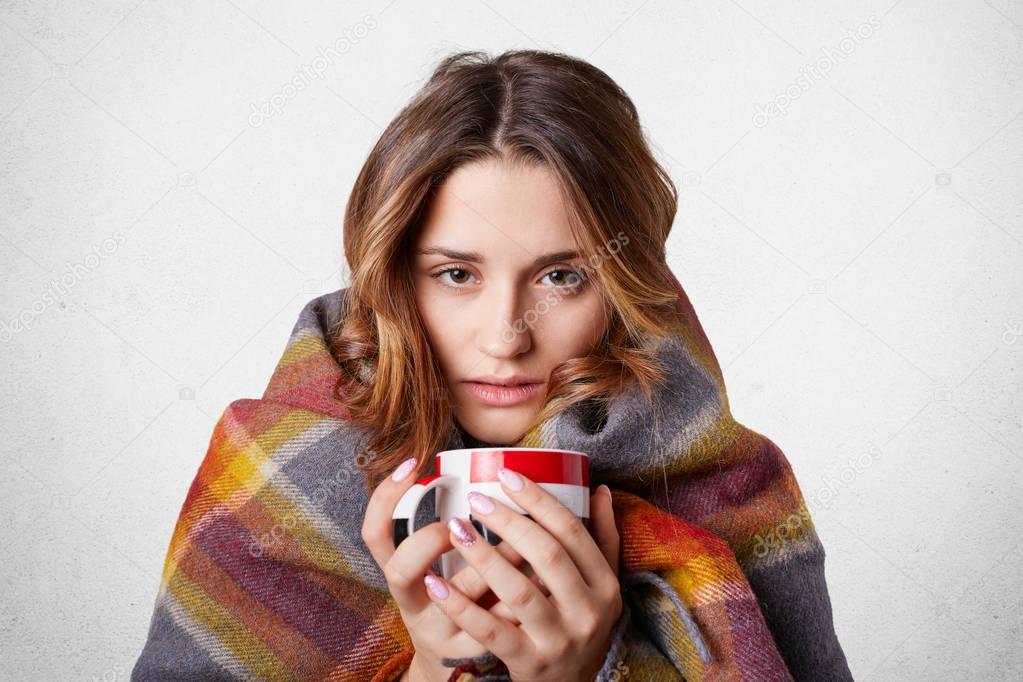 Winter cold sickness concept. Freezing beautiful woman wrapped in warm checkered plaid blanket, drinks hot beverage, tries to warm herself after spending time outside, home warm cozy atmosphere