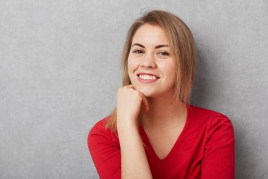 Hoş kadın vardır memnun seyir hoş gülümseme, mükemmel dişler beyaz gösterir, el popüler dergide fotoğraflandı, gri arka plan üzerinde izole memnun olmak çene altında tutar.