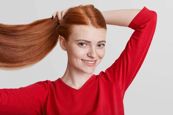 Retrato de pelirroja feliz bonita joven mujer demuestra su lujoso pelo largo, hace cola de caballo, va a tener paseo con amigos, mira directamente a la cámara, posa sobre fondo blanco . — Foto de Stock