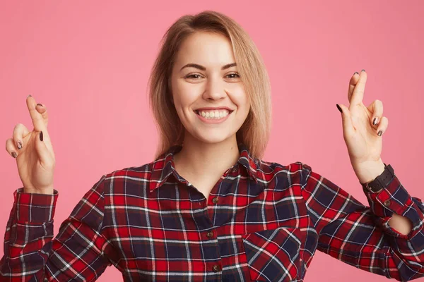 Mujer sonriente europea vestida con camisa a cuadros, cruza los dedos como desea, espera tener buena suerte en el examen, aislado sobre fondo rosa. La mujer cree en su victoria y éxito — Foto de Stock