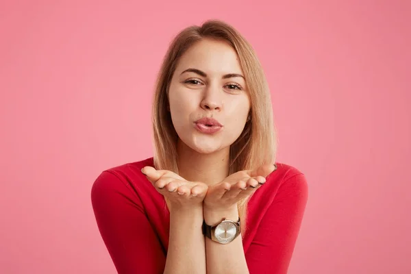 Ritratto di donna positiva indossa maglione rosso e orologio alla moda, soffia bacio come flirta con qualcuno, esprime amore e buoni sentimenti, isolato su sfondo rosa. Glamour donna rende bacio . — Foto Stock