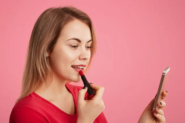 Sideways portrait of female lady paints lips with red lipstick, uses smart phone as mirror, prepares for date with handsome man, applies cosmetic, isolated over pink background. Beauty concept