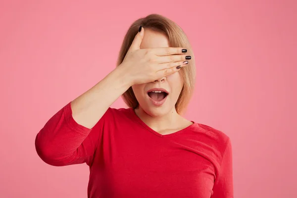 Mujer bonita cubre su cara con la mano, abre la boca, trata de esconderse de alguien, vestido con ropa casual, aislado sobre fondo rosa. Mujer europea siendo sorprendido, cubre los ojos espera sorpresa — Foto de Stock