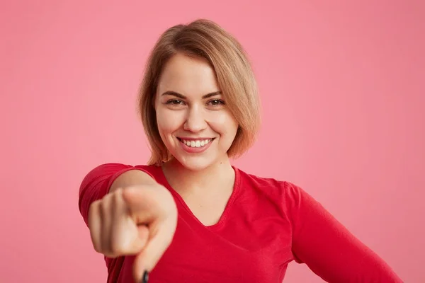 Sorrindo muito jovem fêmea tem amplo sorriso brilhante indica para você com o dedo indicador, faz a escolha, feliz de ser fotografado, isolado sobre fundo rosa. Expressões faciais e gestos — Fotografia de Stock