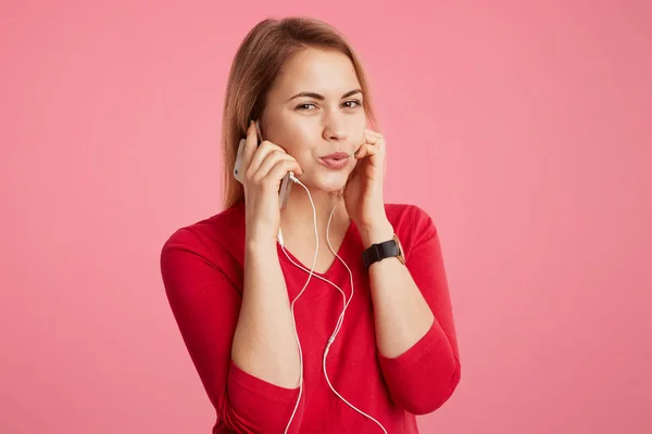 आनंदी महिला इयरफोन्समध्ये आवडत्या ट्रॅकचा आनंद घेतात, आधुनिक गॅझेट वापरतात, गुलाबी पार्श्वभूमीवर वेगळे असलेले आनंददायी संगीत ऐकून आनंदित होतात. लाल स्वेटरमध्ये सुंदर तरुण स्त्री एकटे संगीत ऐकते — स्टॉक फोटो, इमेज
