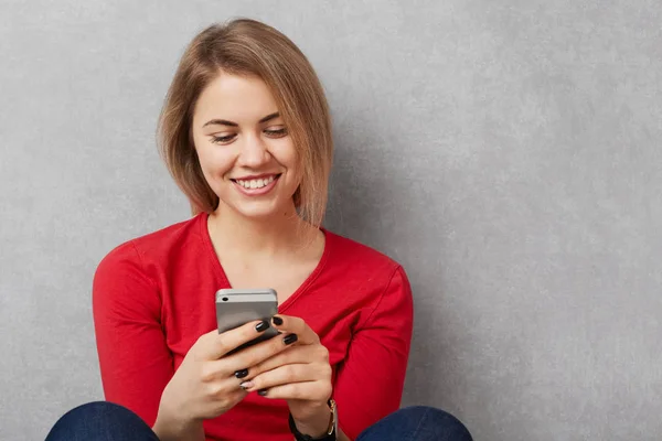 Joyeux sourire jeune femme en pull rouge tient téléphone intelligent et messages en ligne, heureux de recevoir un message texte, assis sur le sol, isolé sur fond gris avec de l'espace de copie. Concept technologique — Photo