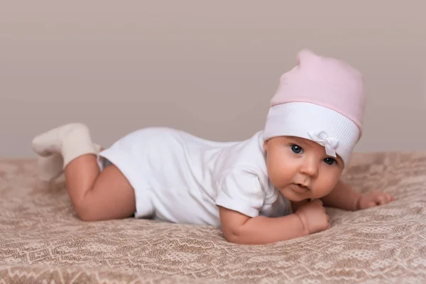 Linda menina recém-nascida se arrasta na cama, vestida com belo chapéu rosa, olha inocentemente para a câmera. Pequeno bebê gosta de tempo com os pais. Uma filha gira no quarto. Conceito de infância e beleza — Fotografia de Stock