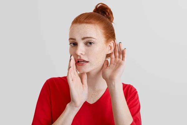 Hermosa mujer foxy trata de escuchar o escuchar a escondidas chismes de alguien, mantiene la mano cerca de la oreja, se ve con secreto, aislado sobre fondo blanco. Curiosa pelirroja joven mujer escucha algo — Foto de Stock