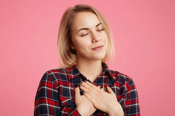 Ramah baik hati perempuan terus mata tertutup, tangan di dada, mengungkapkan rasa terima kasihnya kepada seseorang, berpose terhadap latar belakang merah muda. Wanita muda cantik yang manis santai di dalam ruangan, menjadi cantik — Stok Foto