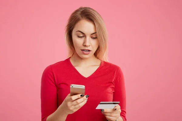 Indoor shot of puzzled female doesn`t recieve salary, checks bank account via online application, uses modern cell phone and plastic card, types password, isolated on pink background. Online shopping — Stock Photo, Image