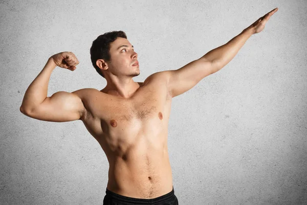 Foto eines fitten, kräftigen jungen männlichen Bodybuilders posiert, zeigt angespannte Muskeln, streckt Hände, isoliert vor grauem Hintergrund. Fitnessmodel mit nacktem Oberkörper demonstriert seine Stärke und seinen sportlichen Körper — Stockfoto