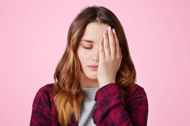 Isolated shot of pretty frustrated overworked female covers face with hand, keeps eyes shut, being sleepy, feels bored as spends time alone, poses against pink background. Tiredness concept. clipart