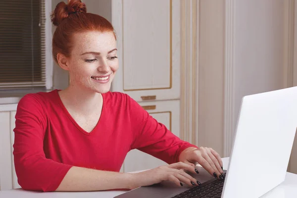 Ευτυχής φακιδωμένος γυναίκα ντυμένη στα κόκκινα πουλόβερ πληκτρολόγια πληροφορίες στον φορητό υπολογιστή, κάθεται πάνω από την κουζίνα, εσωτερικό, αναζητά πληροφορίες online. Χαρούμενα σπουδαστής δουλεύει σε notebook μετά διαλέξεις — Φωτογραφία Αρχείου