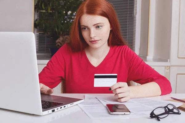 Serious freckled ginger female uses laptop computer for e commerce, holds plastic card, checks balance, going to recieve salary soon. Young female freelancer registers on online bank to get money