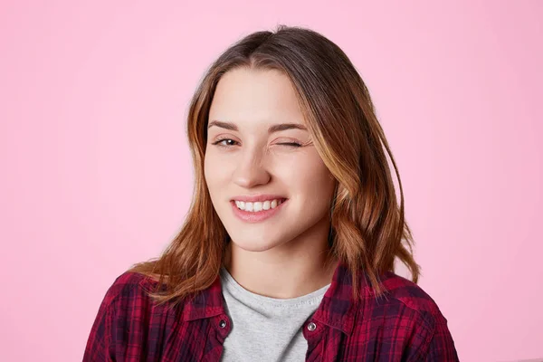 Retrato de mujer con expresión alegre, parpadea mientras coquetea con un hombre guapo, expresa su simpatía, vestida con camisa casual a cuadros rojos, aislada sobre fondo rosa. Mujer guiña un ojo interior — Foto de Stock