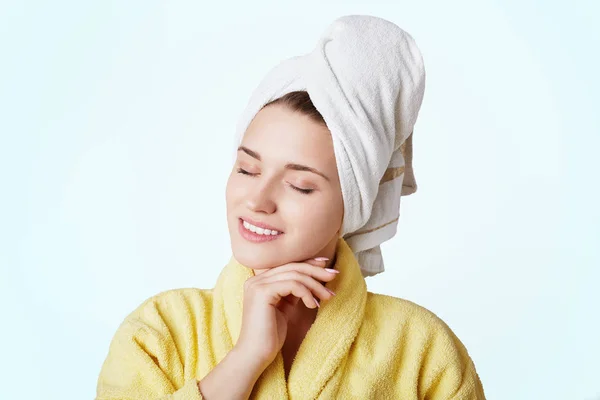 Pleased young female model feels enjoyment after taking shower, has soft skin, wears comfortable gown and towel on head, closes eyes with peasure, isolated over white background. Skin care concept — Stock Photo, Image